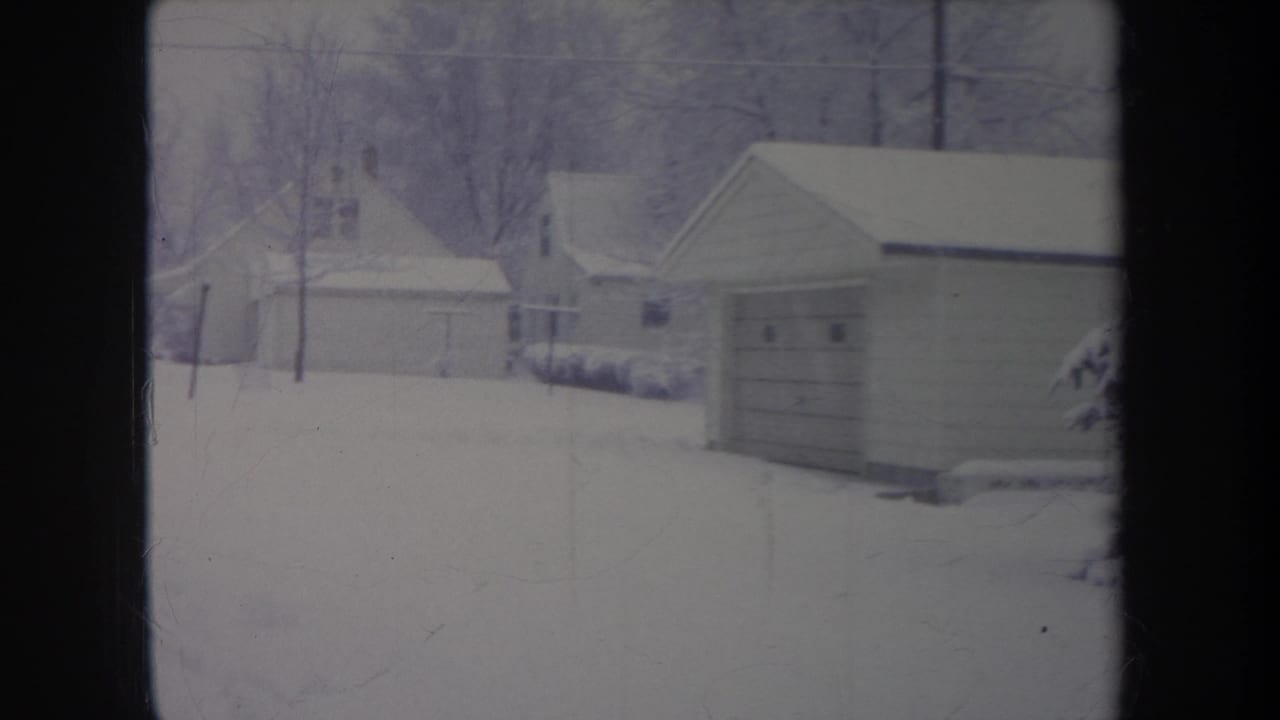 Snow-covered garages and houses seen through a soft, cool-toned home-movie frame.