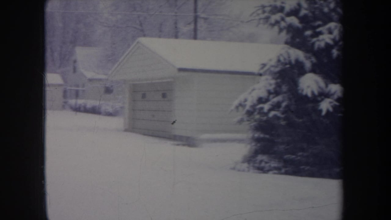 1968 Michigan home-movie frame showing a garage, heavy snow on a pine tree, nearby houses, and a blanketed yard.
