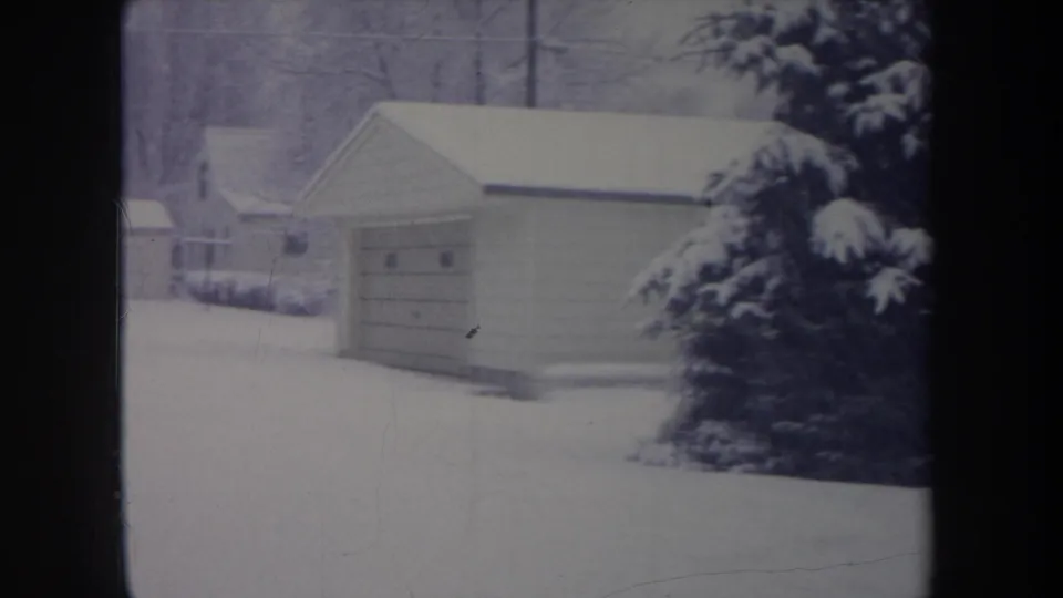 1968 Michigan home-movie frame showing a garage, heavy snow on a pine tree, nearby houses, and a blanketed yard.