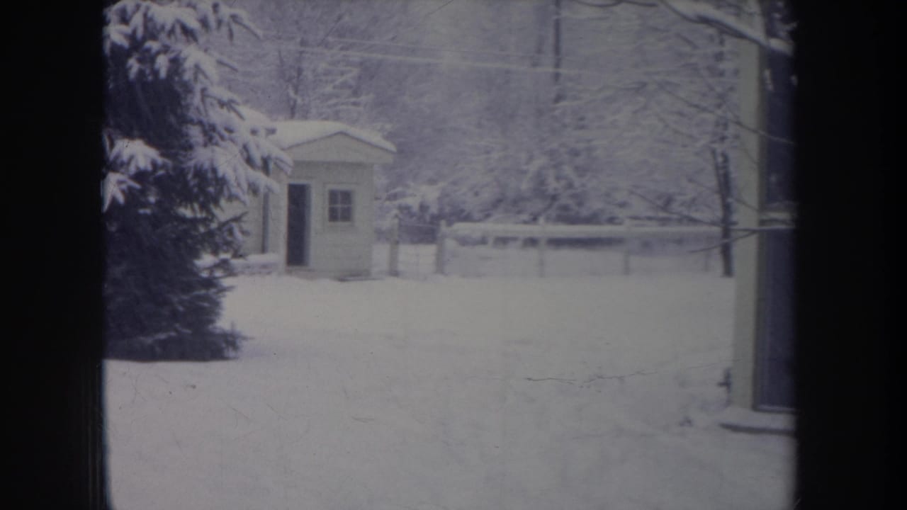 1968 Michigan winter frame showing a small shed beyond deep snow, with evergreen branches, fence, and bare trees.