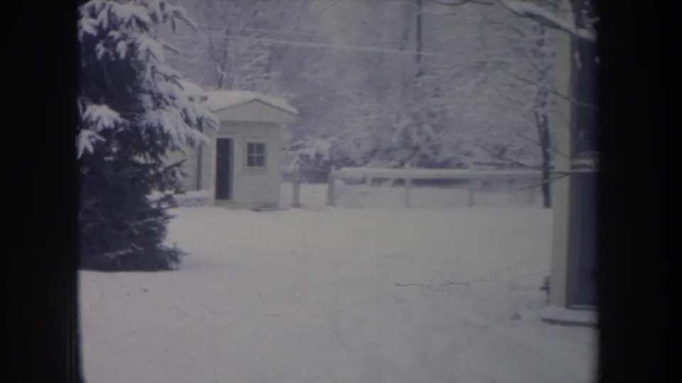 1968 Michigan winter frame showing a small shed beyond deep snow, with evergreen branches, fence, and bare trees.