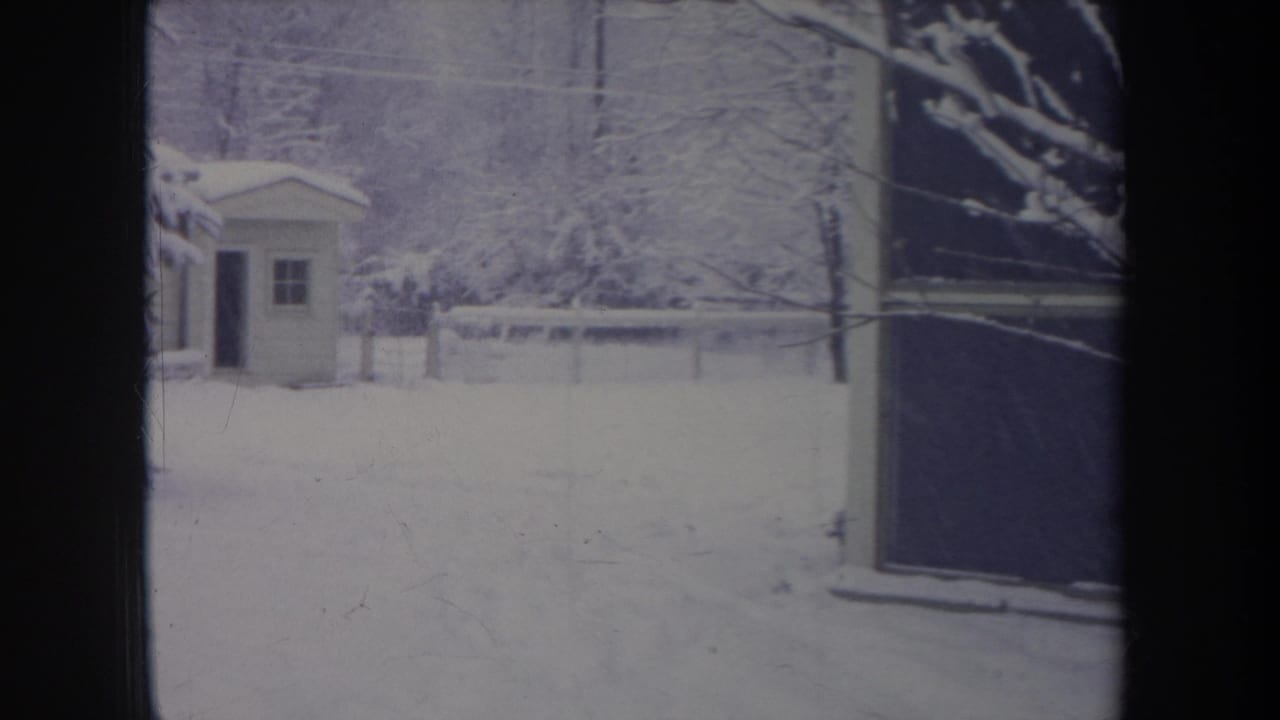 1968 Michigan 8mm frame of a snowy residential yard, small shed, fence, bare trees, and a dark window or doorway edge.