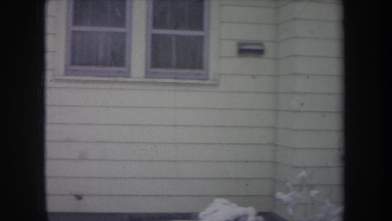 1968 Michigan home-movie frame showing a house wall with paired windows, siding, mailbox, and snow at the base.