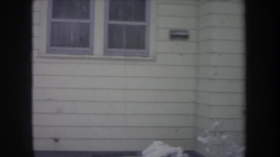 1968 Michigan home-movie frame showing a house wall with paired windows, siding, mailbox, and snow at the base.