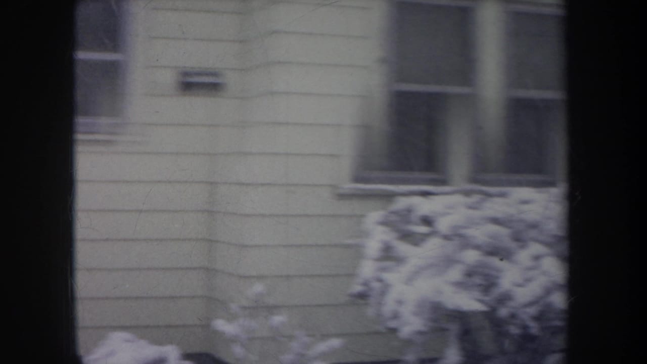 1968 Michigan 8mm frame of a light-colored house exterior with snow piled on shrubs, viewed through dark vertical framing.