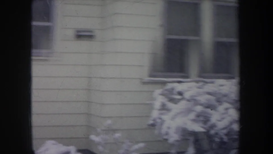 1968 Michigan 8mm frame of a light-colored house exterior with snow piled on shrubs, viewed through dark vertical framing.