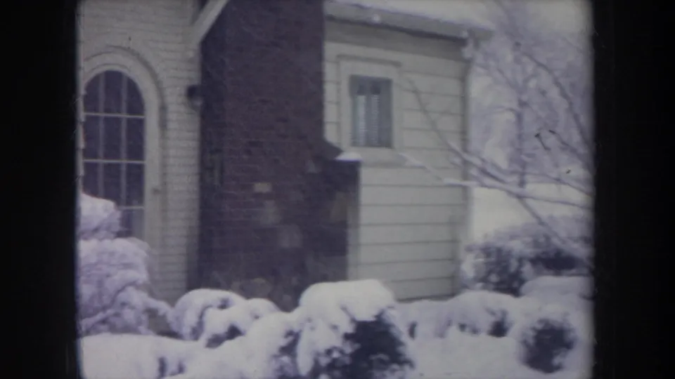 1968 Michigan home-movie view of a brick chimney, arched window, and snow-laden shrubs beside a paneled house.