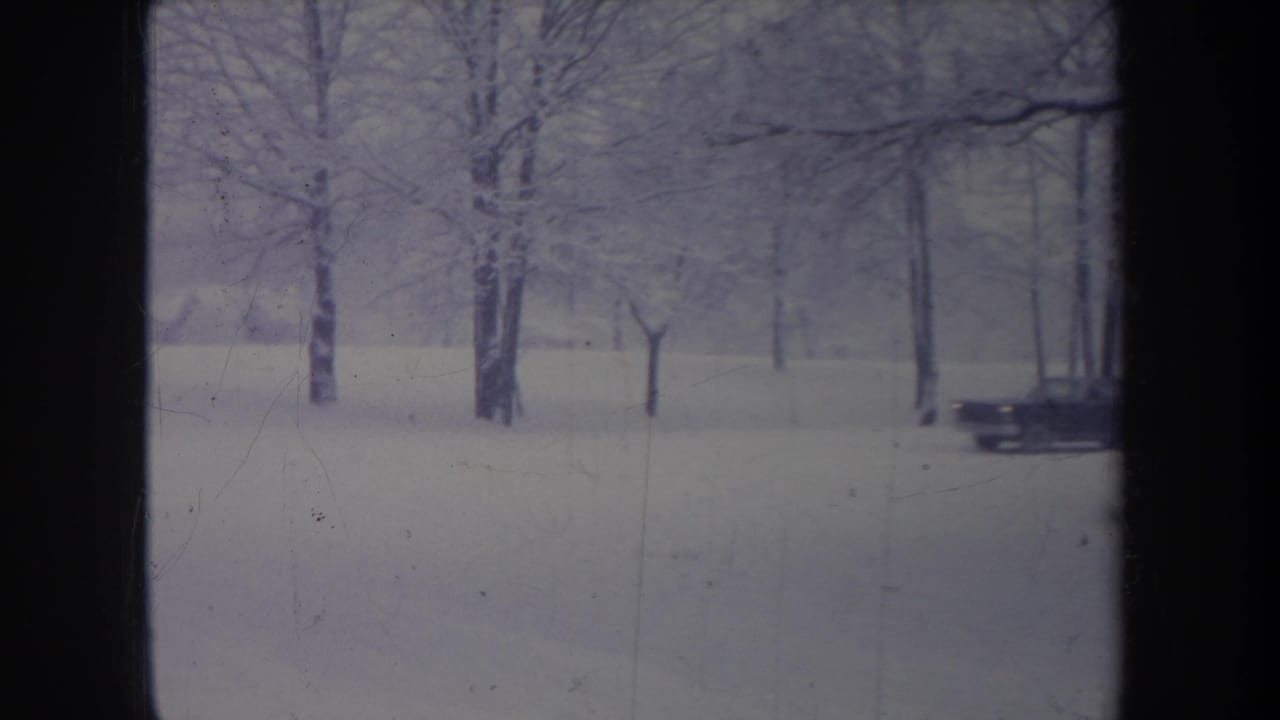 A classic dark sedan drives through a snow-covered Michigan roadside scene, seen through a window-like frame.