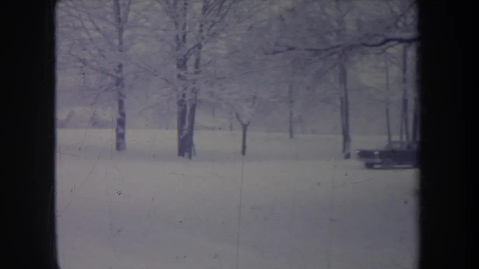 A classic dark sedan drives through a snow-covered Michigan roadside scene, seen through a window-like frame.