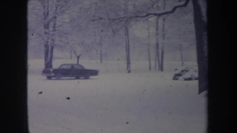 A dark sedan crosses a snowy Michigan road framed by bare trees, with part of another vehicle visible at the edge.
