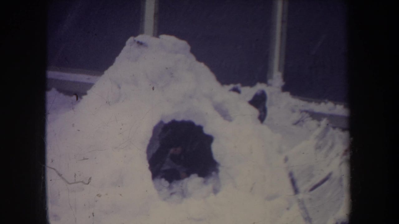 Michigan 1968 home-movie frame showing a handmade snow igloo with a dark entrance beside a structure edge.