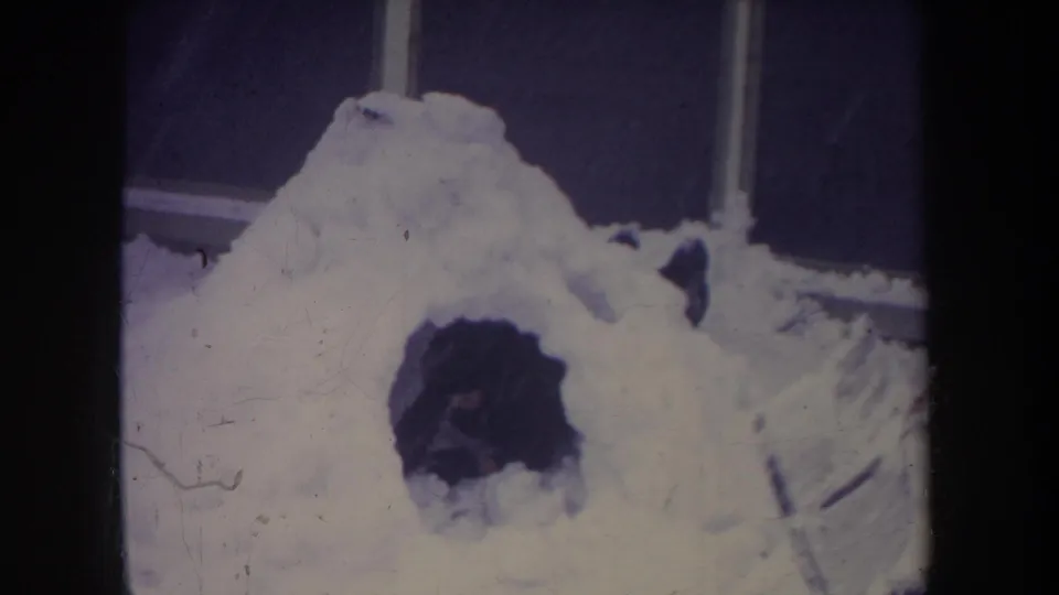 Michigan 1968 home-movie frame showing a handmade snow igloo with a dark entrance beside a structure edge.