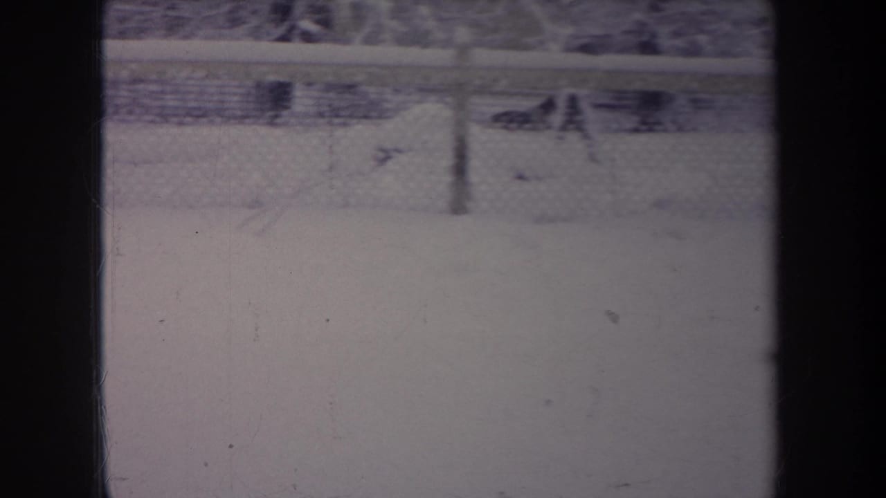 1968 Michigan home-movie frame with snow, a chain-link fence, and dark interior window framing obscuring part of the view.