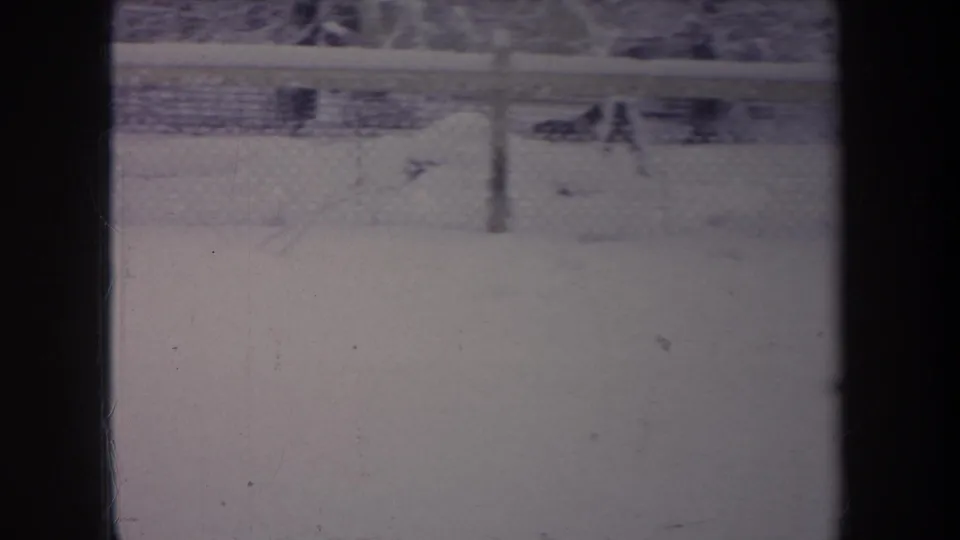 1968 Michigan home-movie frame with snow, a chain-link fence, and dark interior window framing obscuring part of the view.