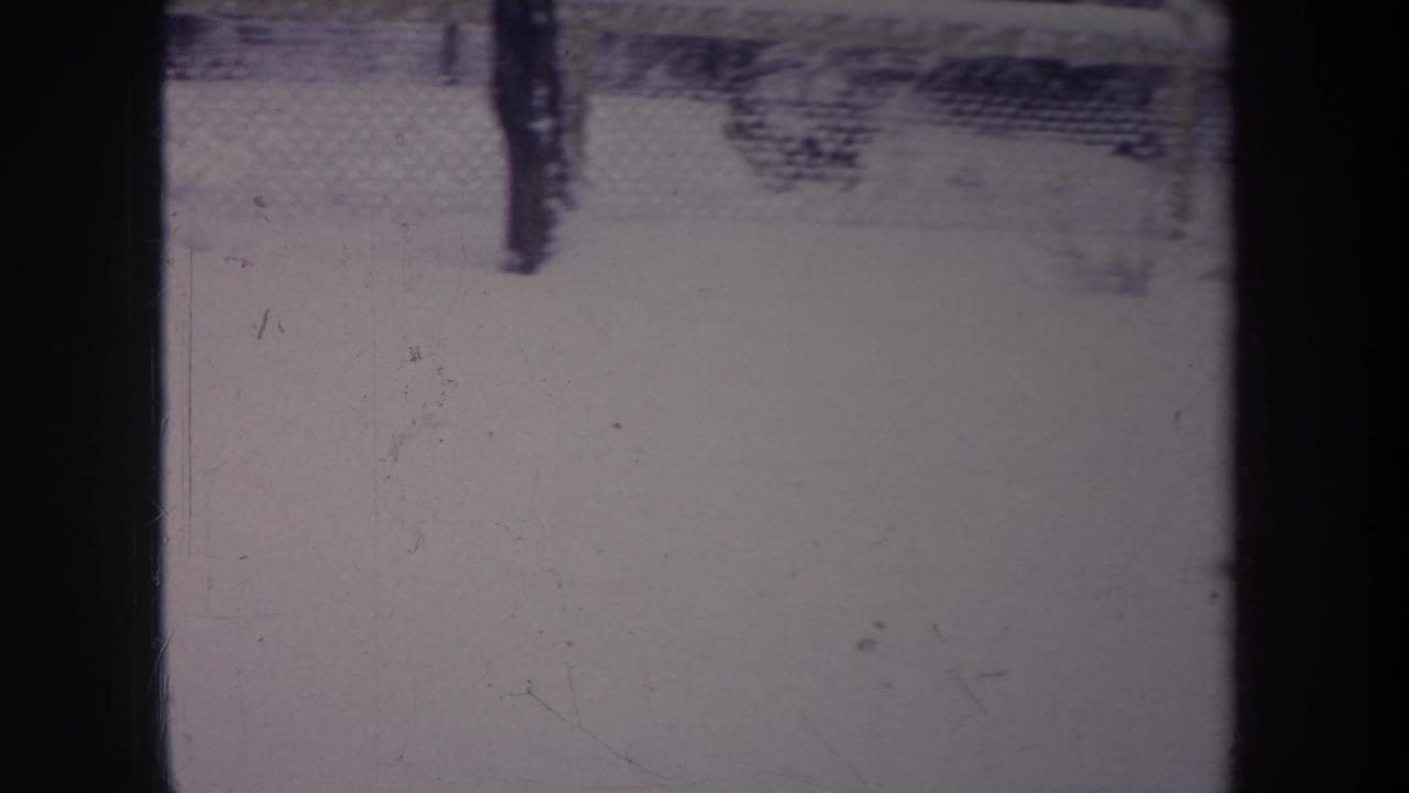 1968 Michigan home-movie frame of a snow-covered backyard viewed past dark vertical bars and a chain-link fence.