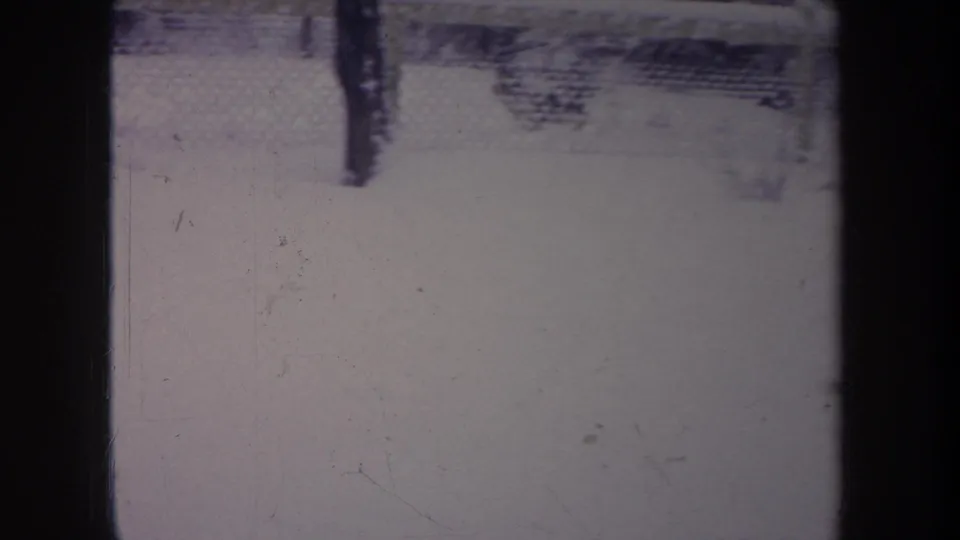 1968 Michigan home-movie frame of a snow-covered backyard viewed past dark vertical bars and a chain-link fence.