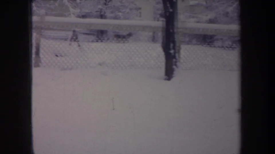 Faded 8mm frame of a snowy Michigan backyard with chain-link fencing and a dark vertical form near the fence.