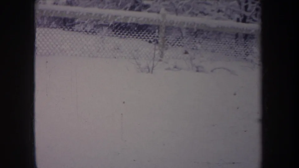 1968 Michigan home-movie frame of a quiet snow-covered yard behind a chain-link fence, with faint shrubs and dark frame obstruction.