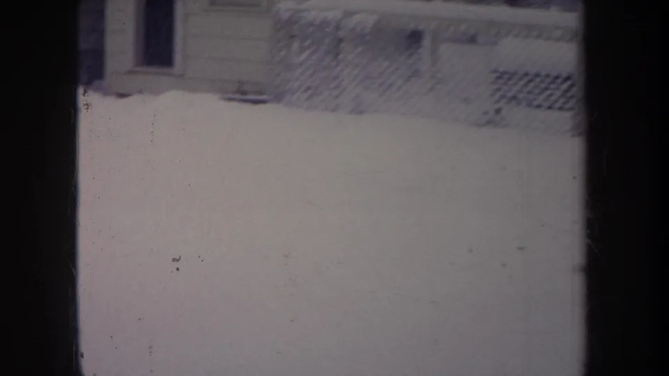 1968 Michigan home-movie frame showing snow beside a chain-link fence and part of a nearby building or yard structure.
