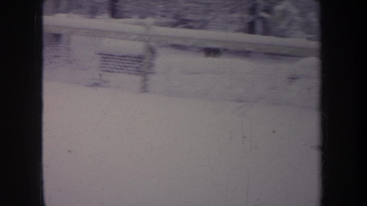 1968 Michigan 8mm frame looking through a window at a snow-covered backyard with a chain-link fence, bare trees, and soft blur.