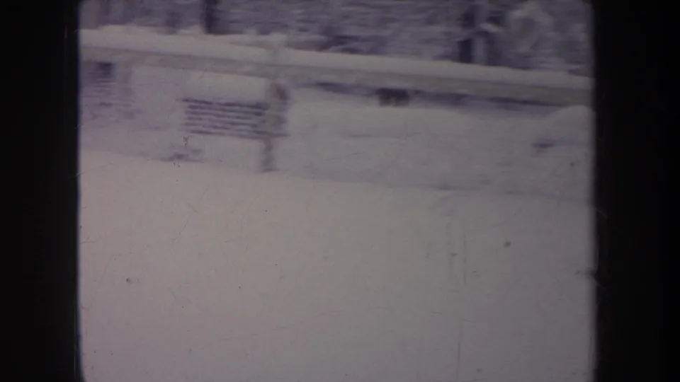 1968 Michigan 8mm frame looking through a window at a snow-covered backyard with a chain-link fence, bare trees, and soft blur.