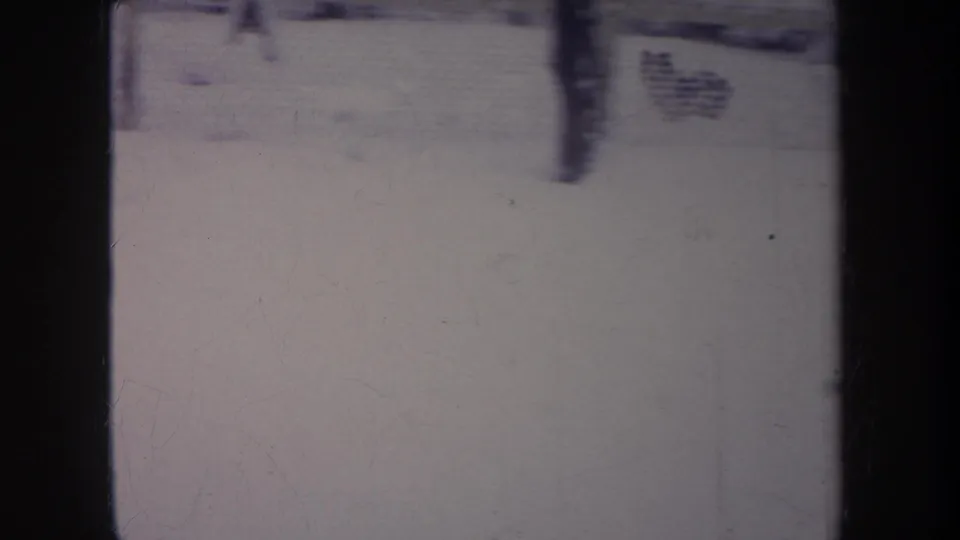 1968 Michigan home-movie frame of a snow-covered backyard seen through window bars, with a fence, tree trunk, and soft purple-tinted film color.