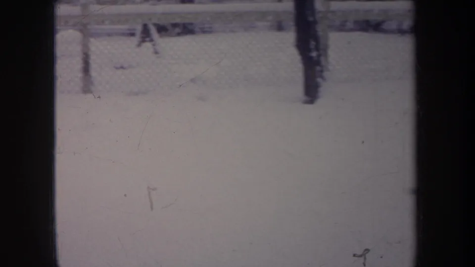 1968 Michigan home-movie frame appearing to show a dark dog near a chain-link fence on snow, with muted color and film marks.