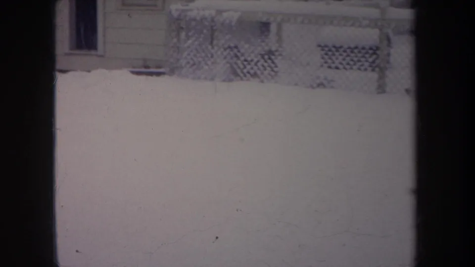 1968 Michigan 8mm backyard frame showing snow, a chain-link fence, and a house exterior with porch lattice in the background.