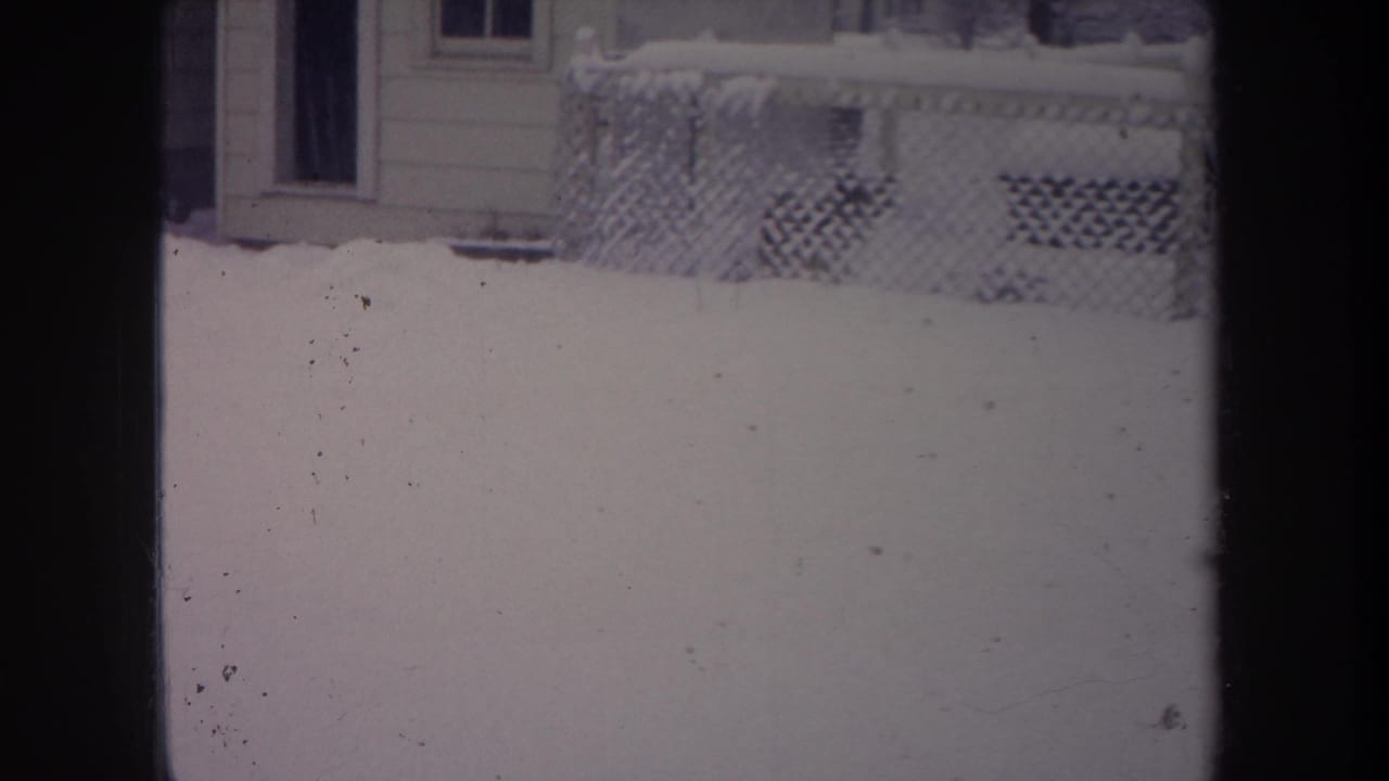 1968 Michigan home-movie frame showing snow outside a window, with a chain-link fence, house siding, porch lattice, and a window visible.