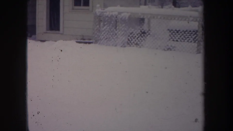 1968 Michigan home-movie frame showing snow outside a window, with a chain-link fence, house siding, porch lattice, and a window visible.