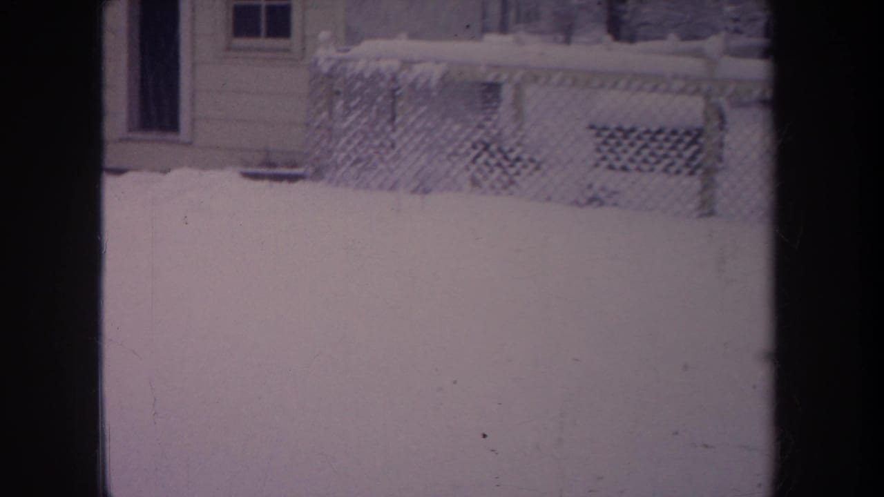 Snow-covered yard and fence viewed through a window, with house siding and porch lattice behind.