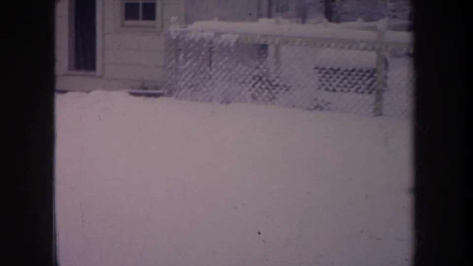 1968 Michigan frame of a snowy backyard through interior window bars, showing a chain-link fence, house wall, porch lattice, and snow piled across the yard.