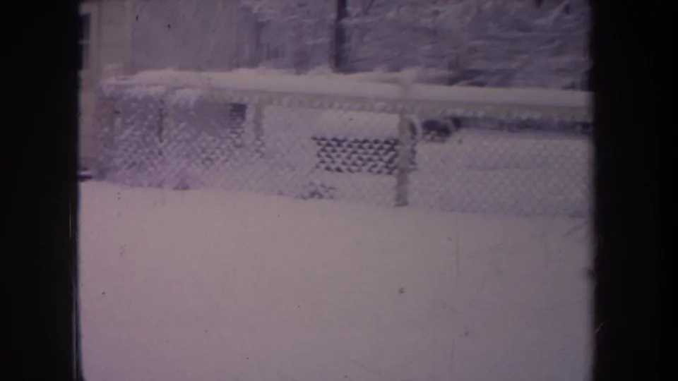 1968 Michigan home-movie still showing a snow-covered residential yard, chain-link fence, porch lattice, and bare trees behind dark window framing.