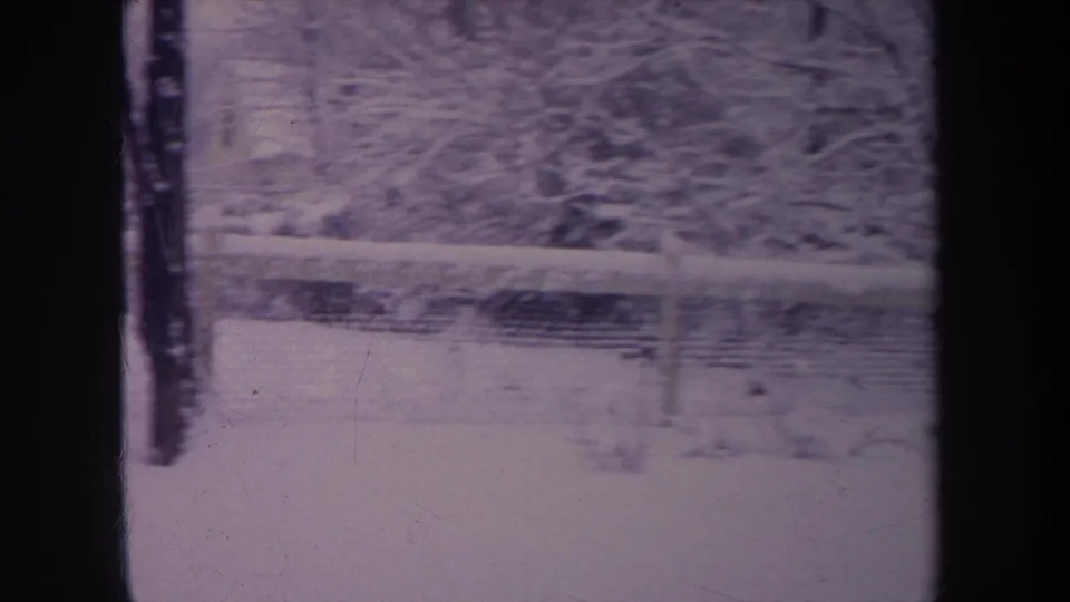 1968 Michigan 8mm frame of a snowy backyard with chain-link fence, bare trees, and dark vertical window or film-frame bars.