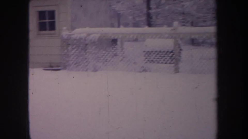 1968 Michigan home-movie frame of a snowy fenced backyard with a house exterior, window, and dark frame bars.