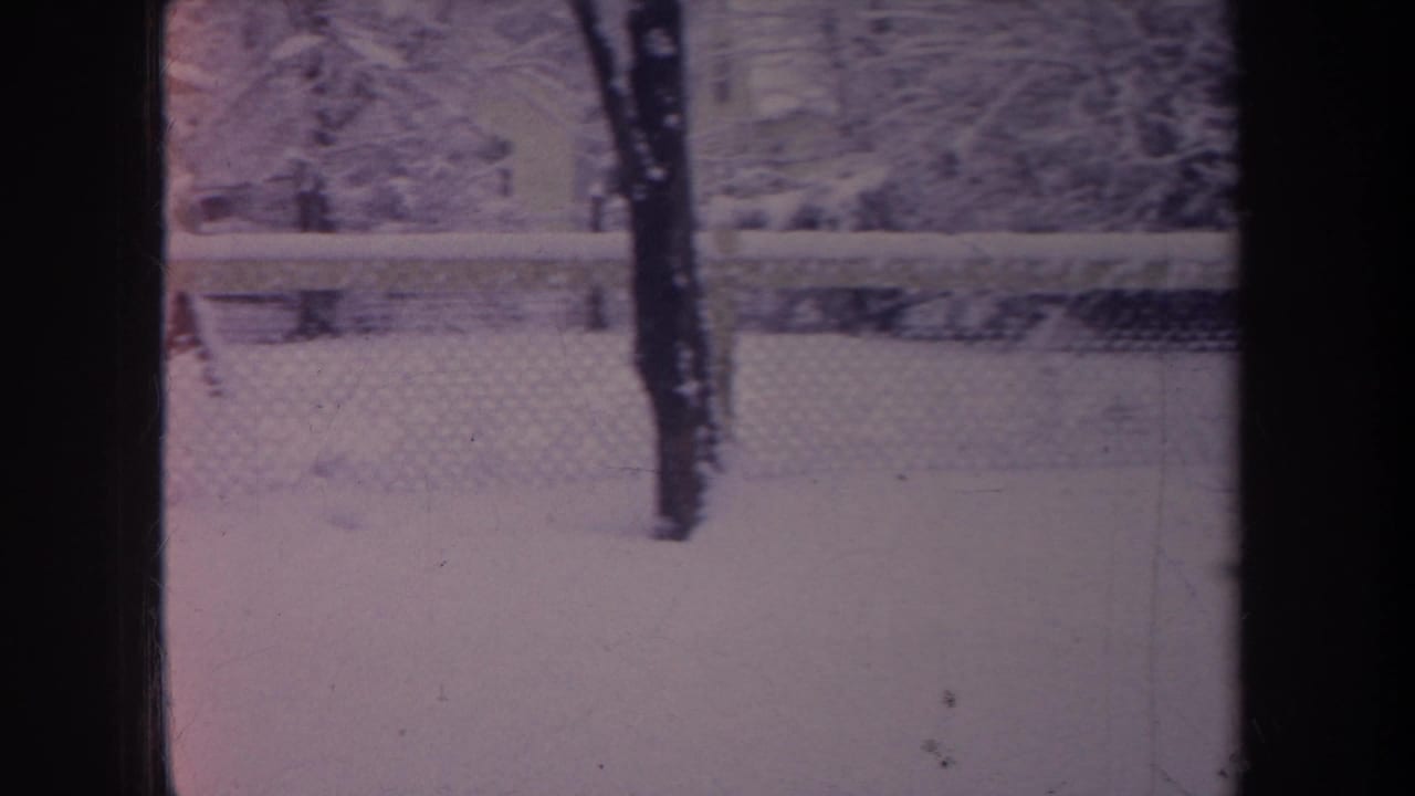 1968 Michigan home-movie frame of snow beyond a chain-link fence, with bare winter trees and dark window framing visible.