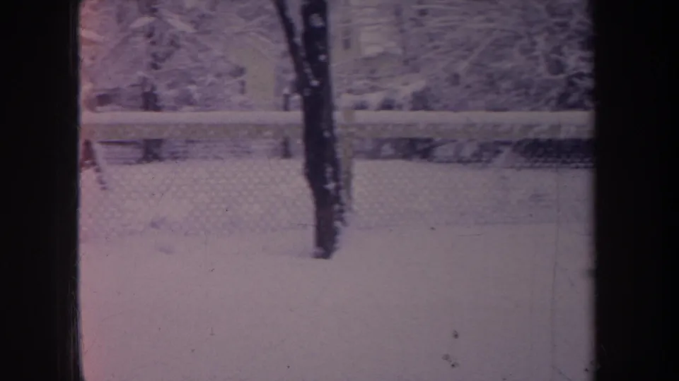 1968 Michigan home-movie frame of snow beyond a chain-link fence, with bare winter trees and dark window framing visible.