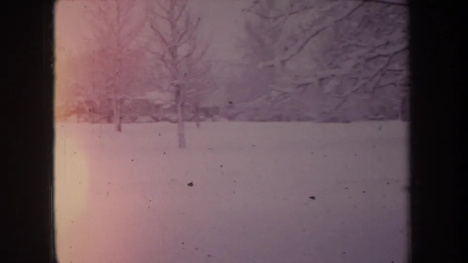 1968 Michigan home-movie frame showing a snowy open area with bare trees, dark window edges, and warm pink-orange film exposure at left.