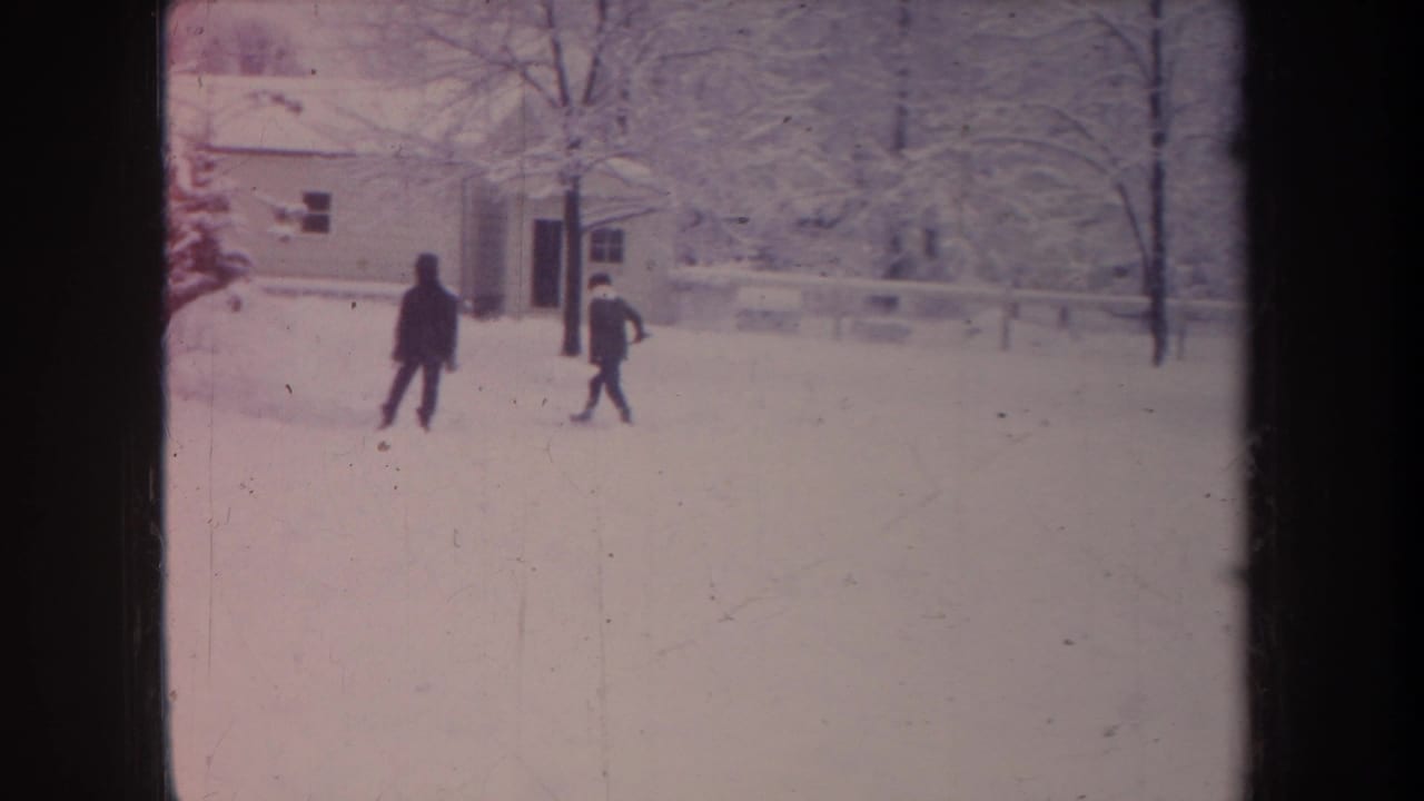 In 1968 Michigan 8mm footage, two distant bundled figures move through a snowy residential yard beside a house and bare winter trees.