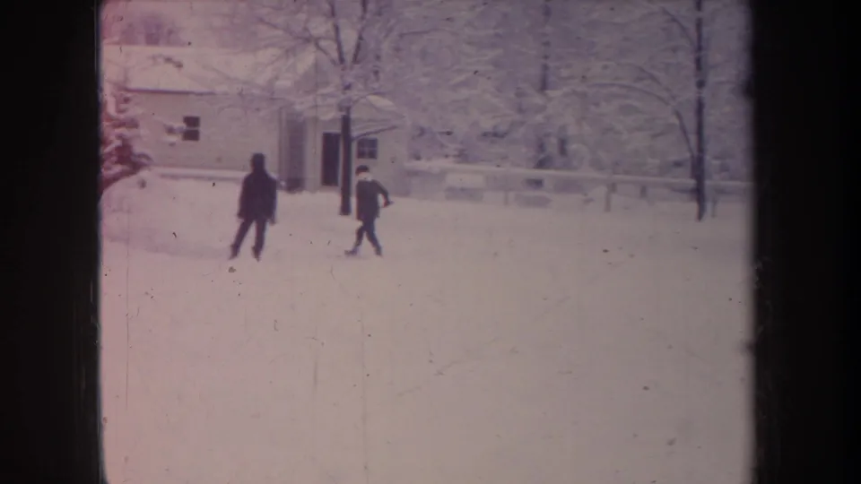 In 1968 Michigan 8mm footage, two distant bundled figures move through a snowy residential yard beside a house and bare winter trees.