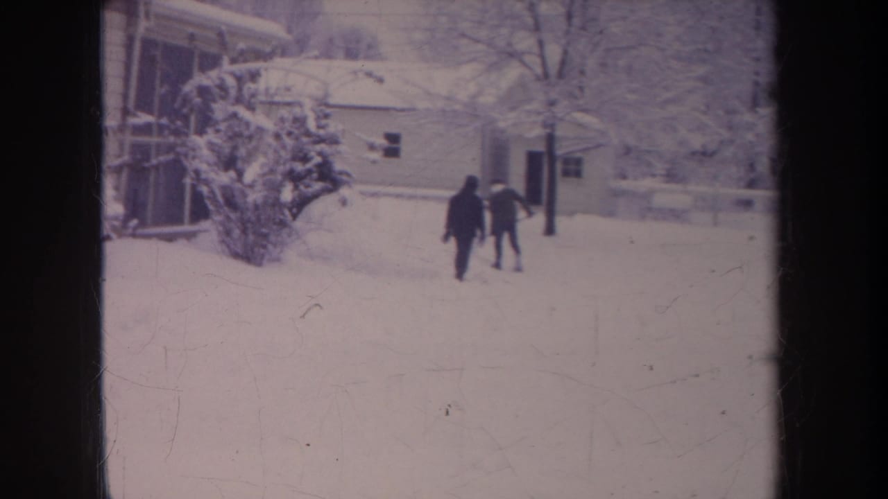 Two bundled figures walk near a snow-covered house and shrub in a winter yard.
