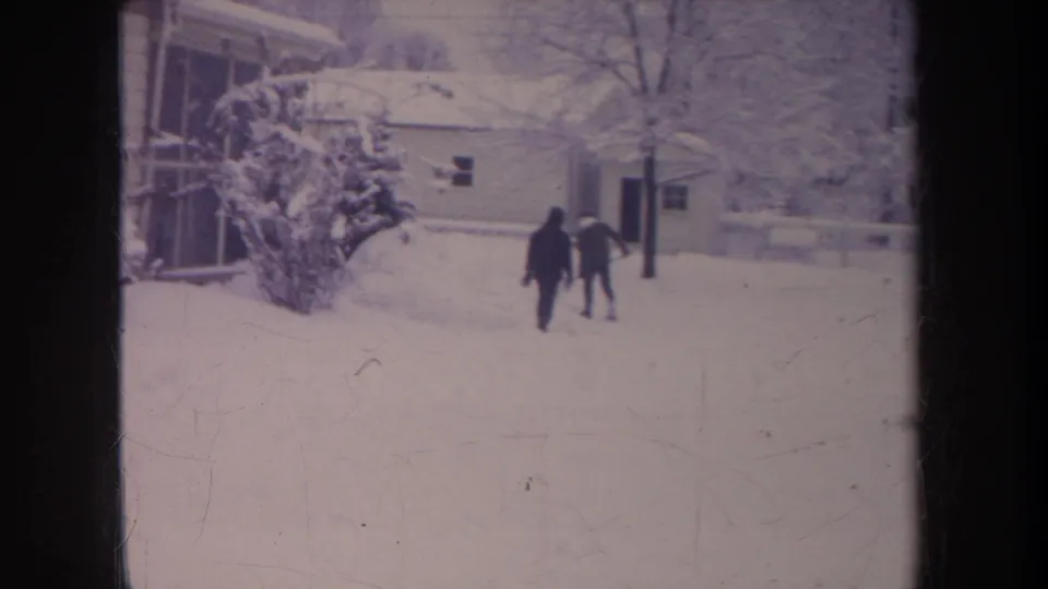 Two distant figures in dark winter clothing walk through deep snow beside a Michigan residential house and snow-laden shrub.