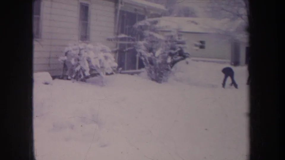 A distant bundled figure bends toward the snow in a residential Michigan yard, with house siding and snow-heavy bushes behind.
