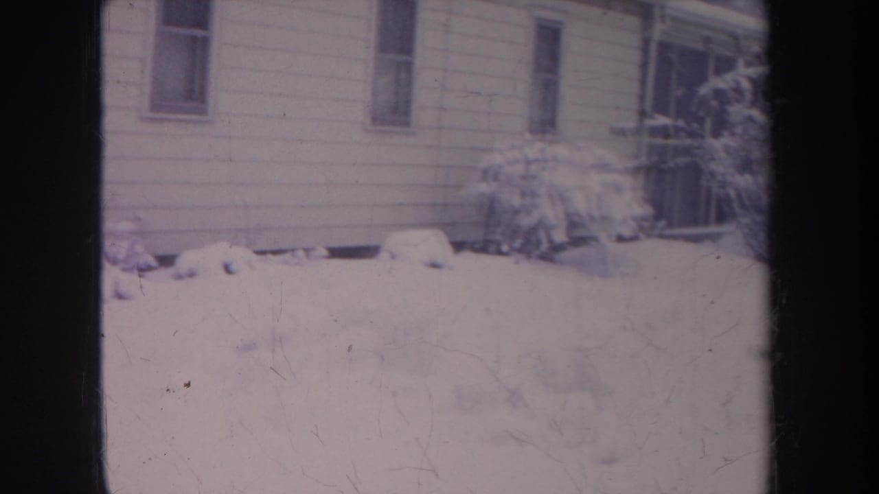 The frame settles on a snowy Michigan residential exterior, showing house siding, narrow windows, bushes, and deep snow in soft 8mm focus.