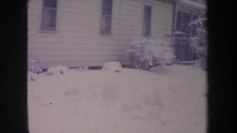 The frame settles on a snowy Michigan residential exterior, showing house siding, narrow windows, bushes, and deep snow in soft 8mm focus.