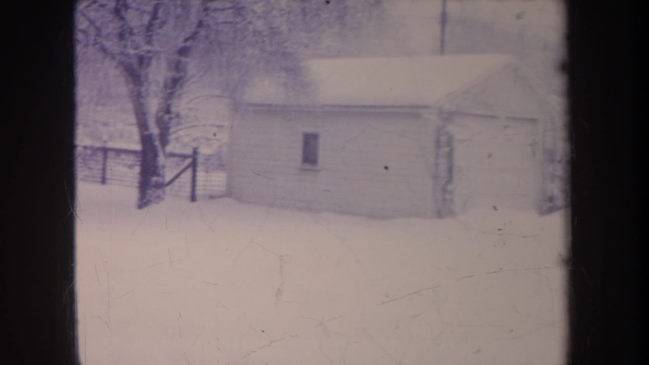 Small garage in deep snow with a tree and fence behind it.