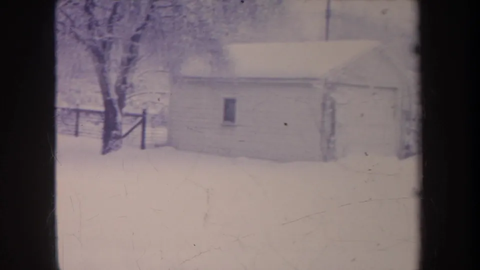 1968 Michigan home-movie frame of a small snow-covered garage, nearby bare tree, and fence line in a quiet winter yard.