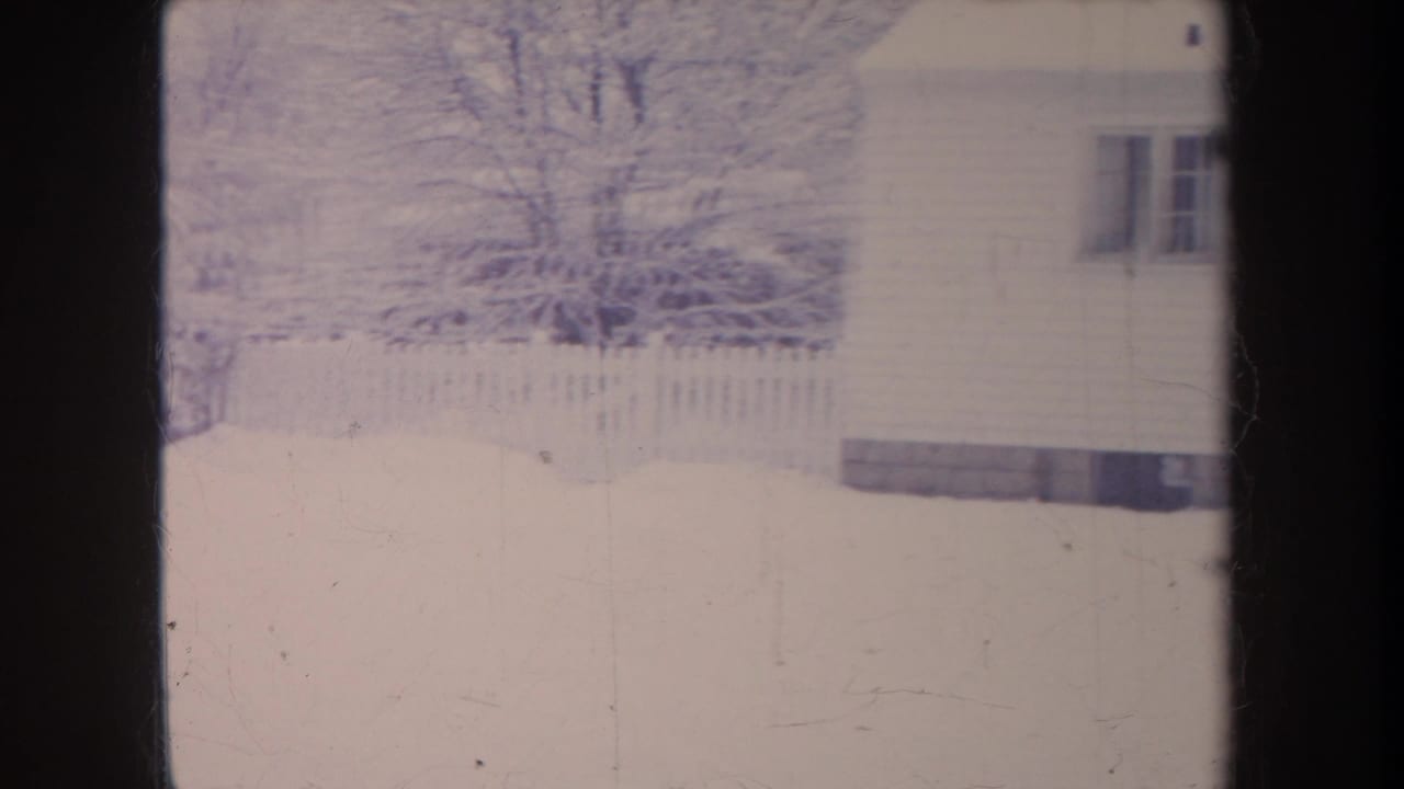 1968 Michigan home-movie frame showing a snow-covered yard, fence, bare trees, and the side of a house at the right edge.