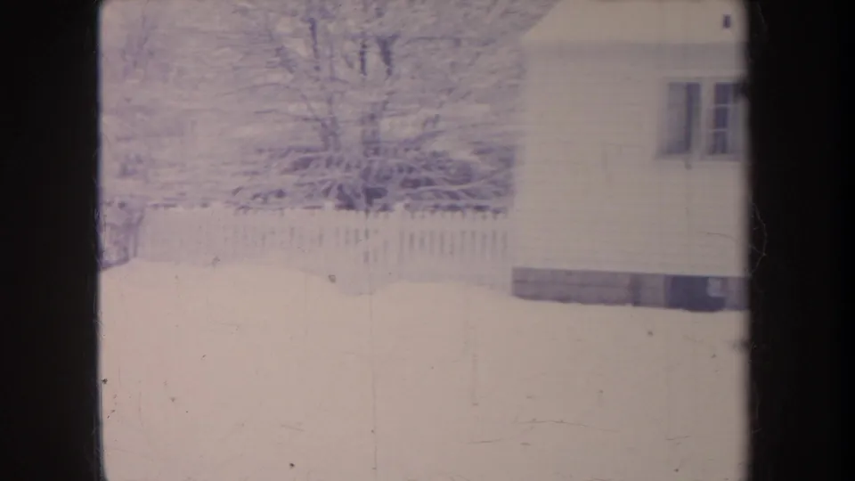 1968 Michigan home-movie frame showing a snow-covered yard, fence, bare trees, and the side of a house at the right edge.