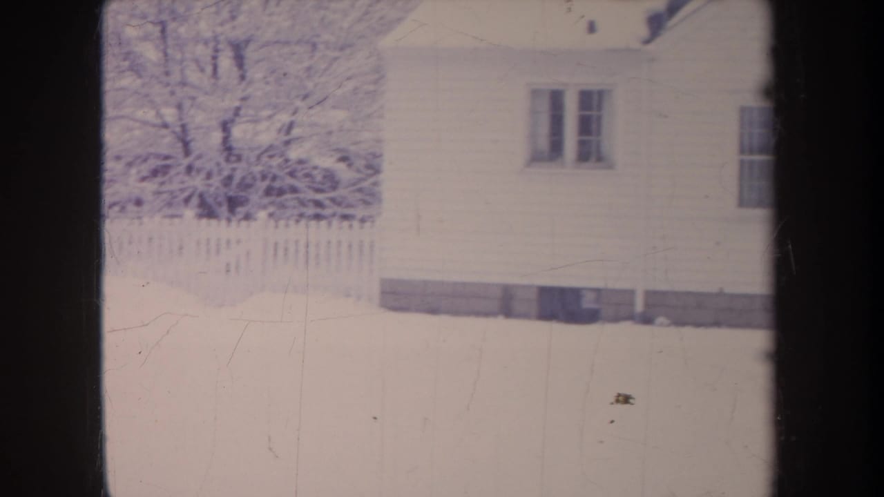 Snowy yard and white clapboard house behind a picket fence.